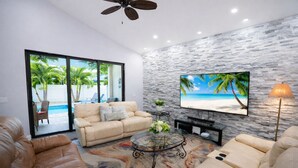 A modern living room with a beige sofa, a glass coffee table, and a television mounted on a stone wall. - Spacious 6bdr/5bath Vacation Home 1 mi to beach (Naples)
