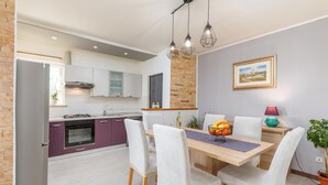 Une cuisine moderne avec des meubles blancs, un îlot de couleur violette et un coin repas comprenant une table en bois et des chaises blanches.