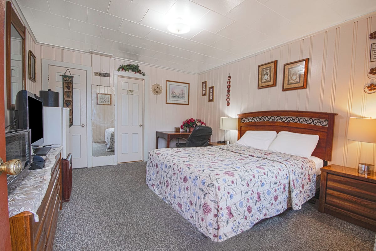 A hotel room with a bed, bedside table, desk, chair, television, and a door leading to another room.