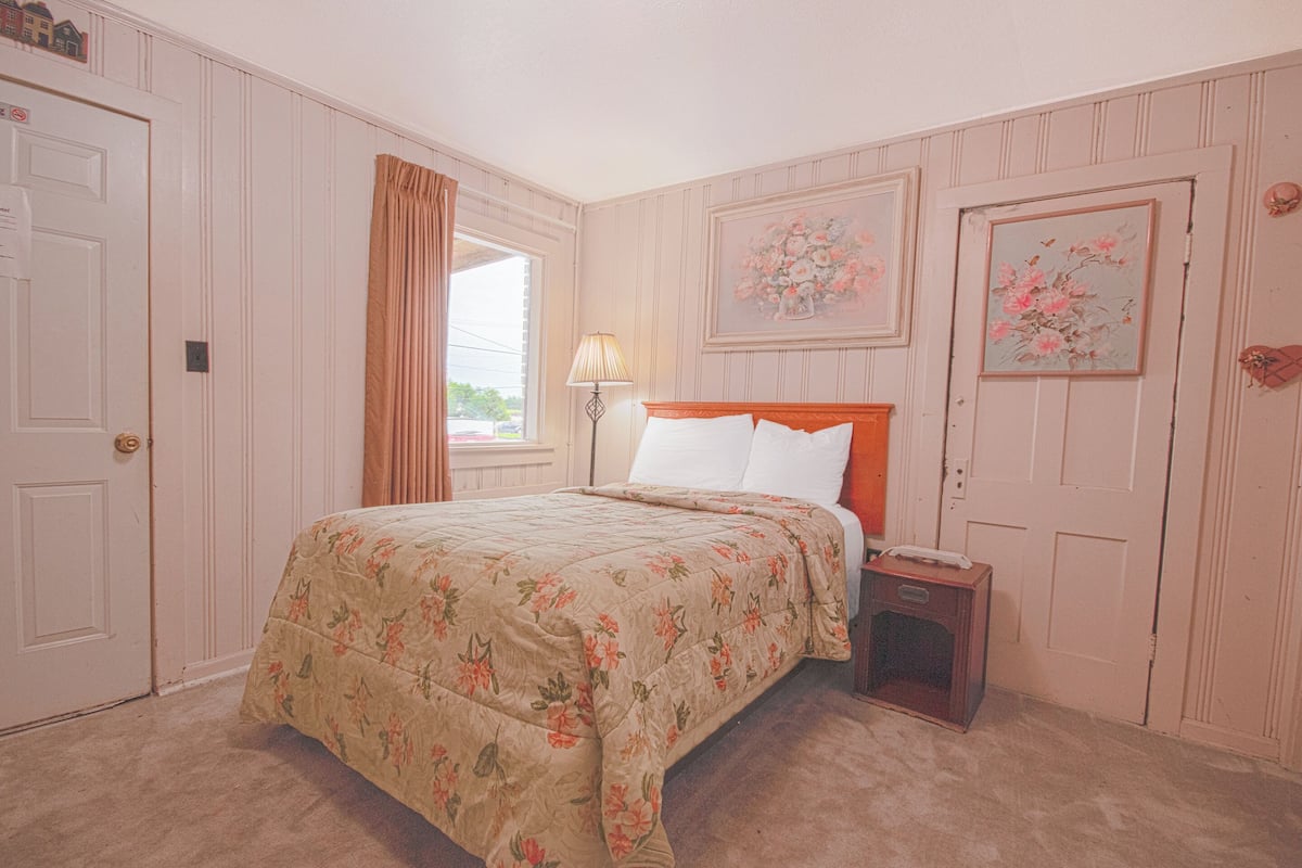 A bedroom with a floral bedspread, a nightstand with a lamp, and a door with a small decorative item.