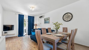 Ein moderner Essbereich mit einem Holztisch, gedeckt für vier Personen, umgeben von Holzstühlen mit blauen Kissen. Eine wandmontierte Uhr und ein gerahmtes Bild sind sichtbar.