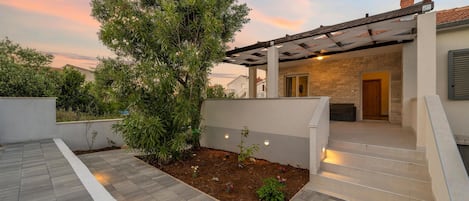 A modern outdoor patio area with a stone wall, a large tree, and a covered seating area.