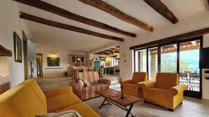 A spacious living area with yellow sofas, a striped armchair, and a wooden coffee table. A kitchen and dining area are visible in the background, with a large window offering a view of the outdoors. - Villa with pool and air conditioning (Bartolići)
