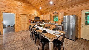 Interior de una cabaña de madera con una mesa de comedor dispuesta para seis personas, un refrigerador de acero inoxidable, armarios de madera y una cocina con microondas y estufa.