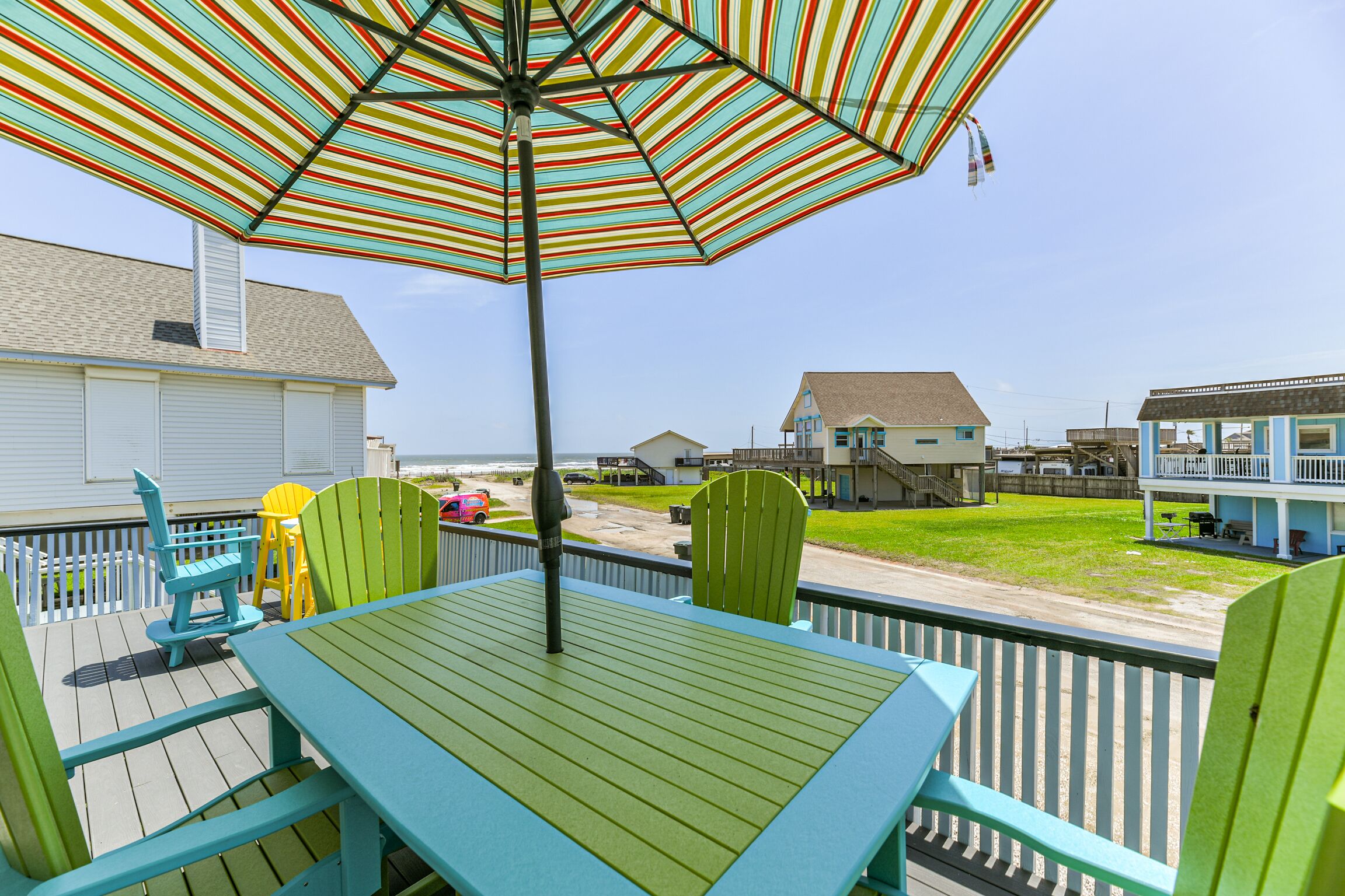 A deck with a table and chairs, an umbrella, and a view of the beach and houses.