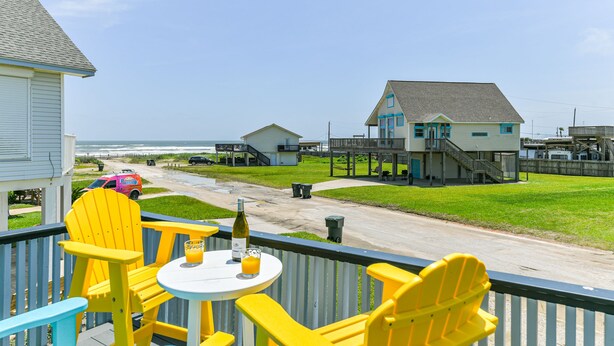 A balcony with yellow chairs and a table, overlooking a beach and a road with houses and a pink car.