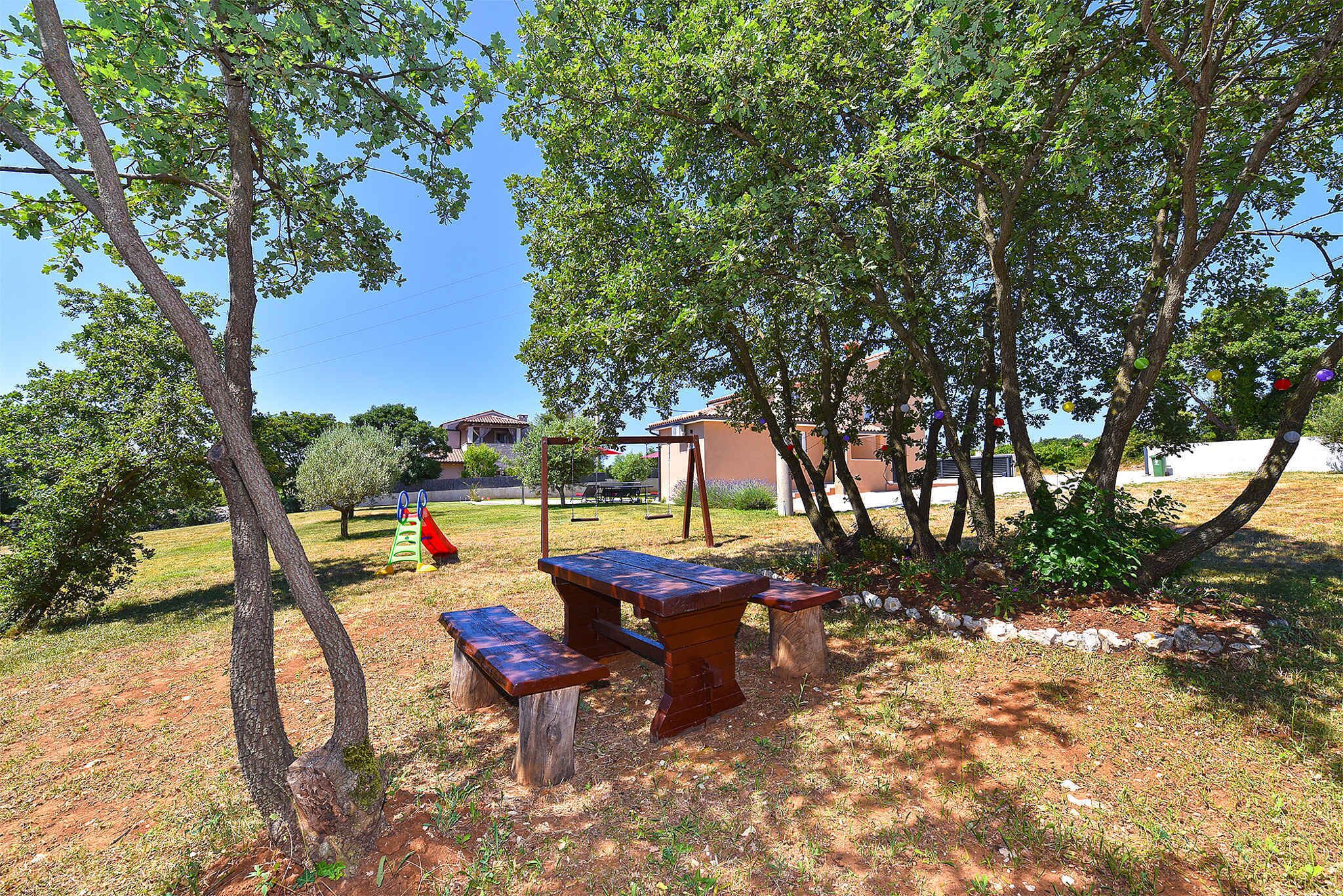 Un espacio al aire libre con mesa y bancos de madera, rodeado de árboles y un campo cubierto de césped.