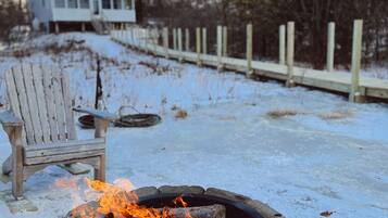 A stone fire pit with flames, a wooden chair, and a snow-covered yard with a wooden fence and a house in the background.