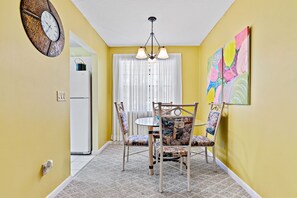 Un coin repas avec une table ronde, des chaises, un lustre, une horloge et un tableau coloré accroché au mur.