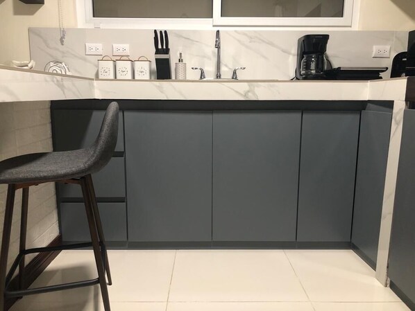 Een keuken met een marmeren aanrecht, grijze kastjes, een barkruk en een koffiezetapparaat.