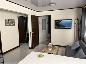 Een moderne woonkamer met een flatscreen televisie aan de muur, een houten salontafel met decoratieve objecten, een bank met kussens en een gang die naar een andere kamer leidt.