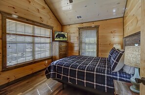 Quarto de cabana de madeira com uma cama, duas janelas com persianas, uma TV fixada na parede e um abajur.