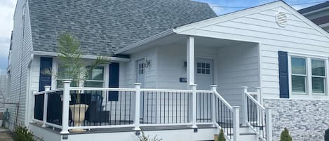 Une maison de plain-pied avec un toit recouvert de bardeaux gris, une véranda blanche dotée d’une balustrade et un petit jardin devant la maison, avec une pelouse et des arbustes.