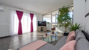 A modern living room with a sofa, a glass coffee table, and a television. There is a large window with curtains, a radiator, and a potted plant. - Gorgeous apartment in Slime (Slime)