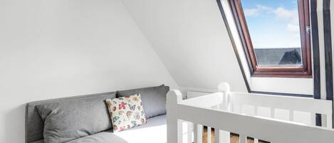 Een kamer met een grijze bank, een bloemenkussen, een witte leuning en een dakraam met uitzicht op een gebouw.