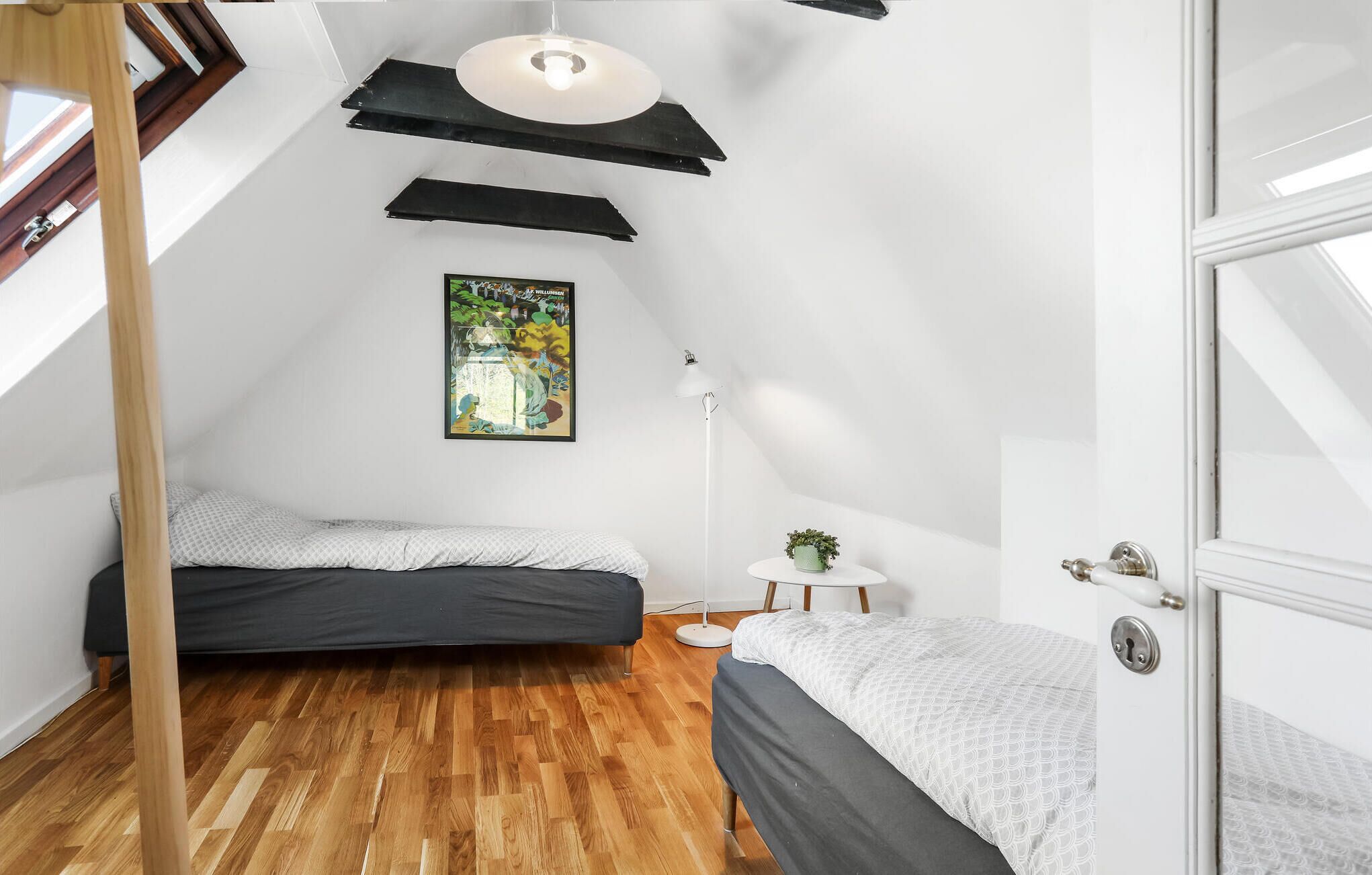 A small, modern bedroom with two beds, a wooden floor, a painting, and a white lamp.