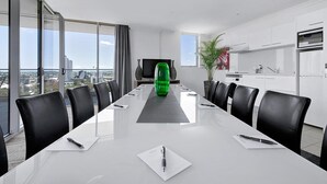 Una sala de conferencias moderna con una mesa larga, sillas negras, un amplio ventanal con vistas a la ciudad y una zona de cocina con armarios blancos.