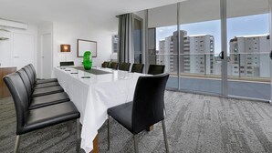 Un salón de conferencias con una larga mesa, sillas, un jarrón verde y vistas a la ciudad a través de amplias ventanas.