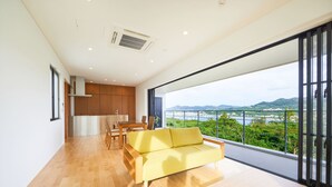 A modern living room with a yellow sofa, wooden flooring, a dining area, and a large window offering a view of greenery and hills.