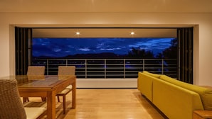 A modern living room with a glass table and chairs, a yellow sofa, and a large window offering a night view of the mountains.