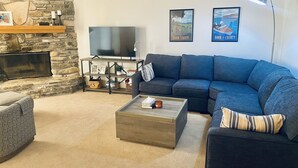 A living room with a stone fireplace, a large flat-screen TV, a blue sectional sofa, a coffee table, and a lamp.