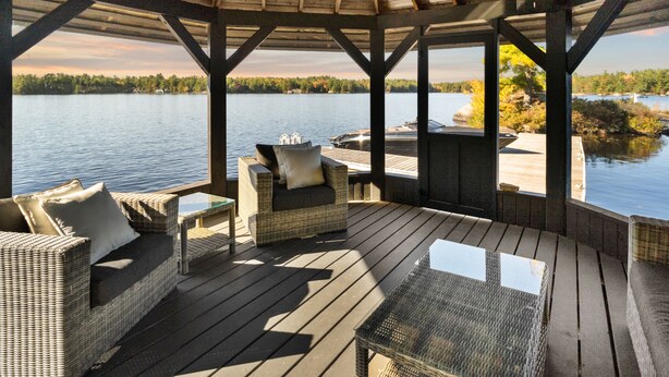 Une terrasse extérieure couverte, aménagée avec des meubles en osier et une table en verre, offrant une vue sur un lac et un quai.