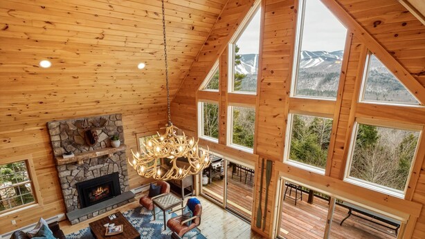 Ein geräumiges Wohnzimmer mit Kamin aus Stein, Holzboden und großen Fenstern mit Blick auf die Berge.