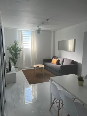 A modern living room with a grey sofa, a coffee table, a dining table with chairs, a ceiling fan, and a large window with curtains. - Arecibo Hotel 3201 (Arecibo)
