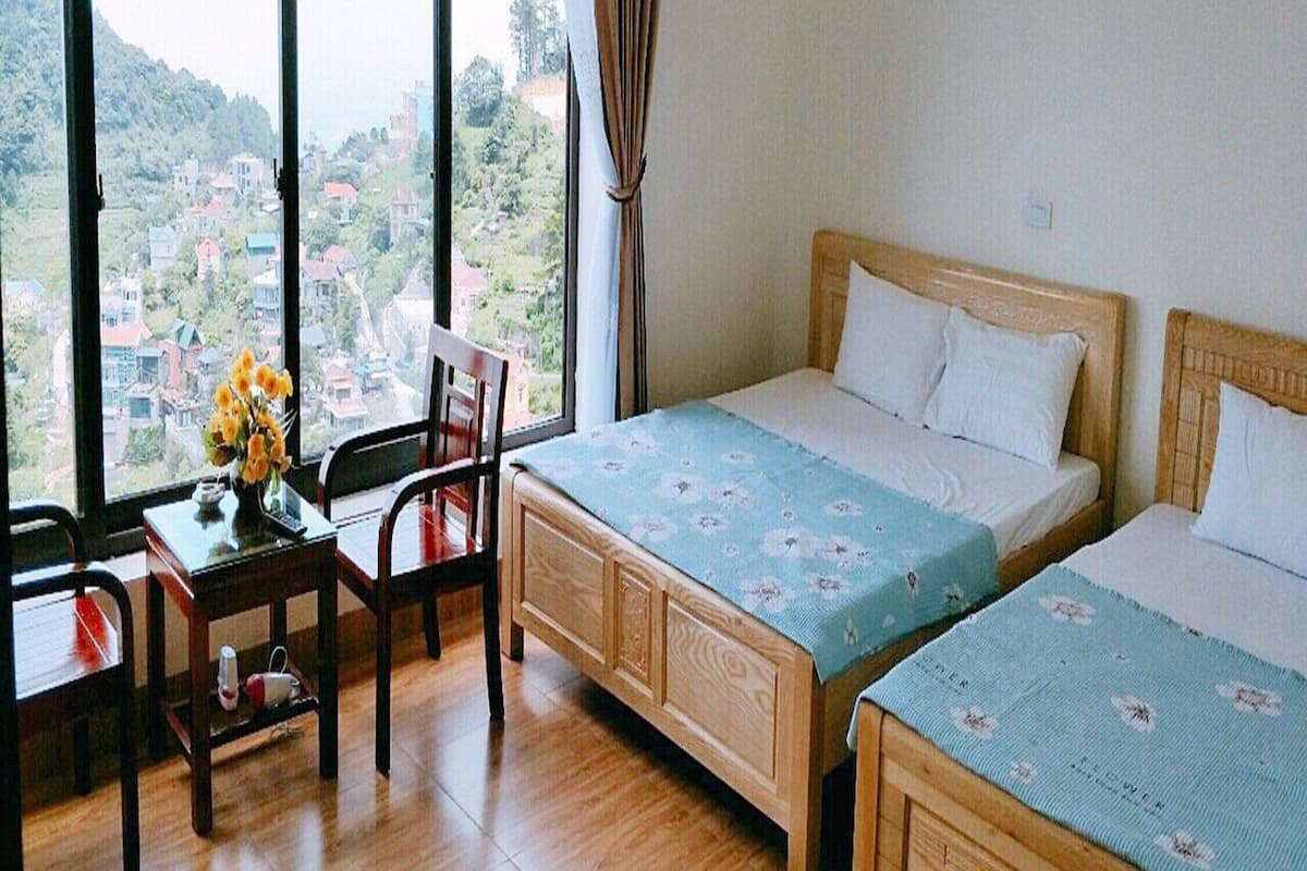 Habitación de hotel con dos camas, un escritorio de madera, una silla y un ventanal que ofrece vistas a un pueblo en la ladera de una colina.