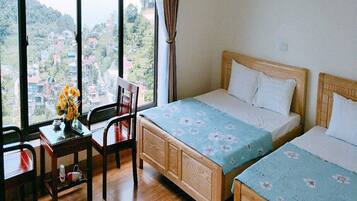 A hotel room with two beds, a wooden desk, a chair, and a large window offering a view of a hillside town.