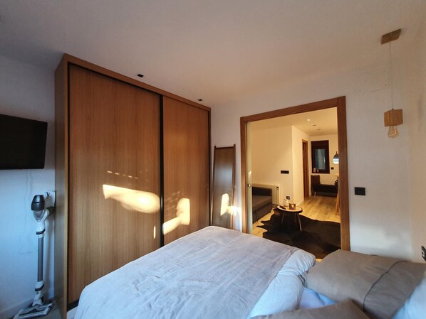 A bedroom with a bed, a wooden wardrobe, a mirror, and a hallway leading to a dining area.