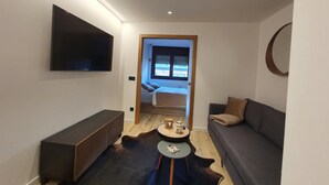 A modern living room with a sofa, a coffee table, a TV mounted on the wall, and a view through an open door to a bedroom.
