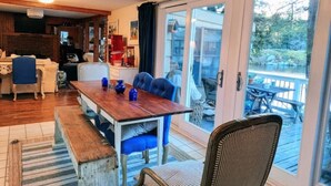 Zona pranzo con tavolo in legno e sedie blu, cucina sullo sfondo e vista su un pontile e un lago attraverso porte in vetro.