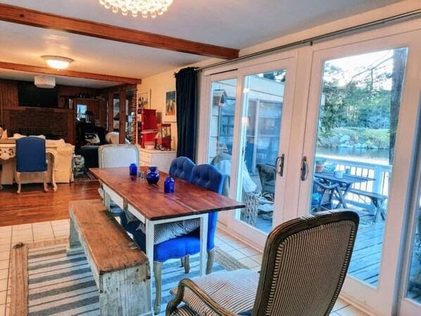 A dining area with a wooden table and blue chairs, a kitchen in the background, and a view of a deck and lake through glass doors. - Cozy Cottage-Sunset On The Lake
(Otis)