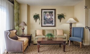 Relax in this inviting living room with a comfortable sofa set, armchair, and coffee table, surrounded by potted plants and a beautiful landscape painting.