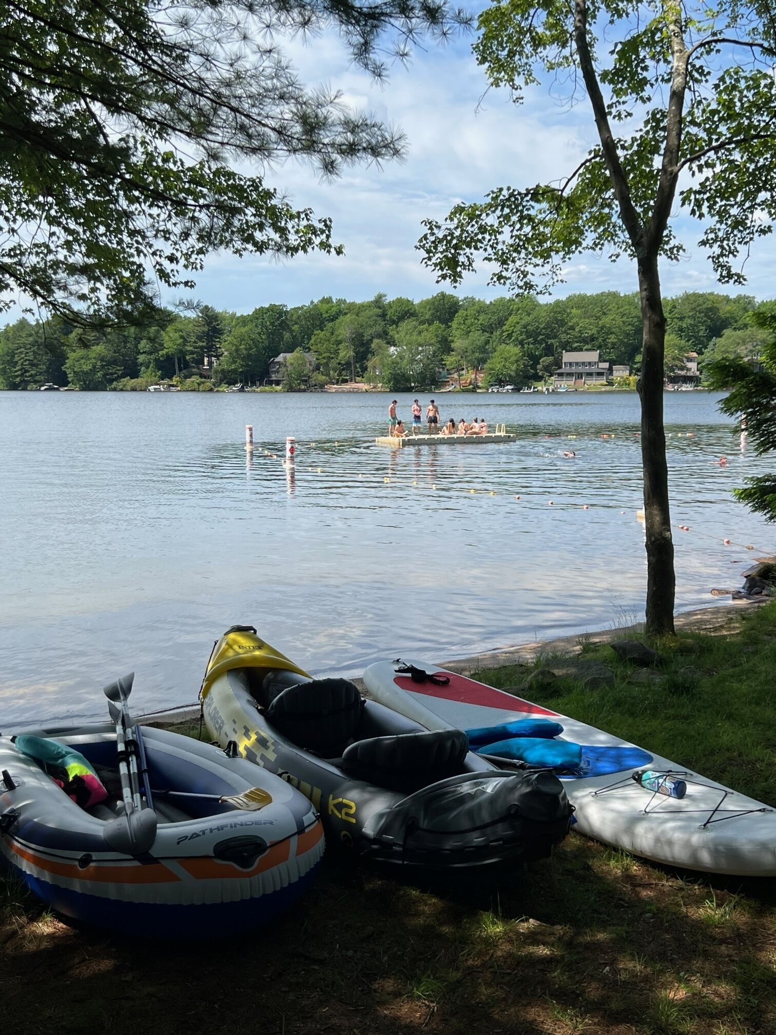 Un paisaje lacustre con kayaks inflables y una tabla de remo, un muelle con gente y un bosque al fondo.