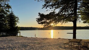 A beach with a wooden bench and a tree, a calm lake, and the sun setting in the background. - Mountain House Hot Tub Pool Table Fire Pit Private beach Walkable to the Lake. (Lake Harmony)