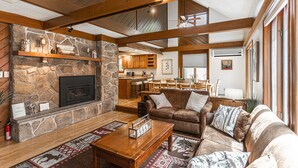 A spacious living room with a stone fireplace, wooden beams, a cozy sofa, and a dining area in the background. - Mountain House Hot Tub Pool Table Fire Pit Private beach Walkable to the Lake. (Lake Harmony)