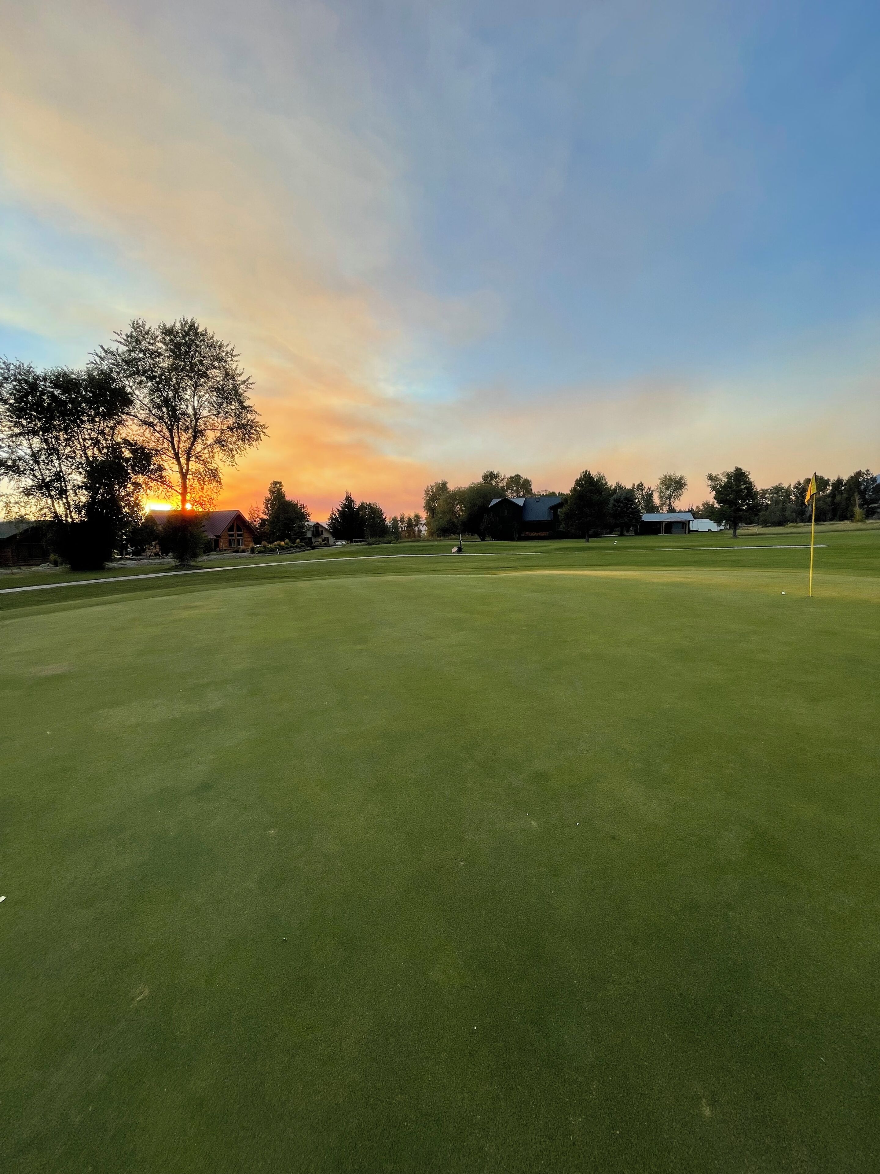 Un campo de golf al atardecer con una bandera, árboles y un edificio en el fondo.