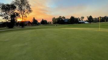 Un campo de golf al atardecer con una bandera, árboles y un edificio en el fondo.