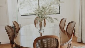 Een eethoek met een ronde houten tafel, zes rieten stoelen, een vaas met takken en een hanglamp.