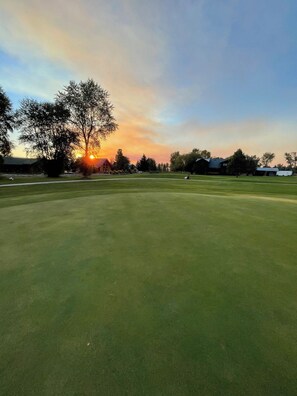 A golf course with a sunset in the background, a large tree, and a building.