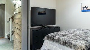 Een slaapkamer met een televisie op een zwarte standaard, een bed met een grijs-witte deken en een houten wand met een afbeelding van een eland.