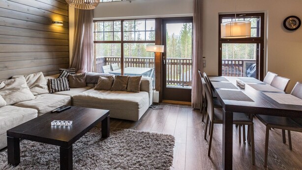 Een ruime woonkamer met een bank, salontafel en eethoek, grote ramen en een houten plafond.