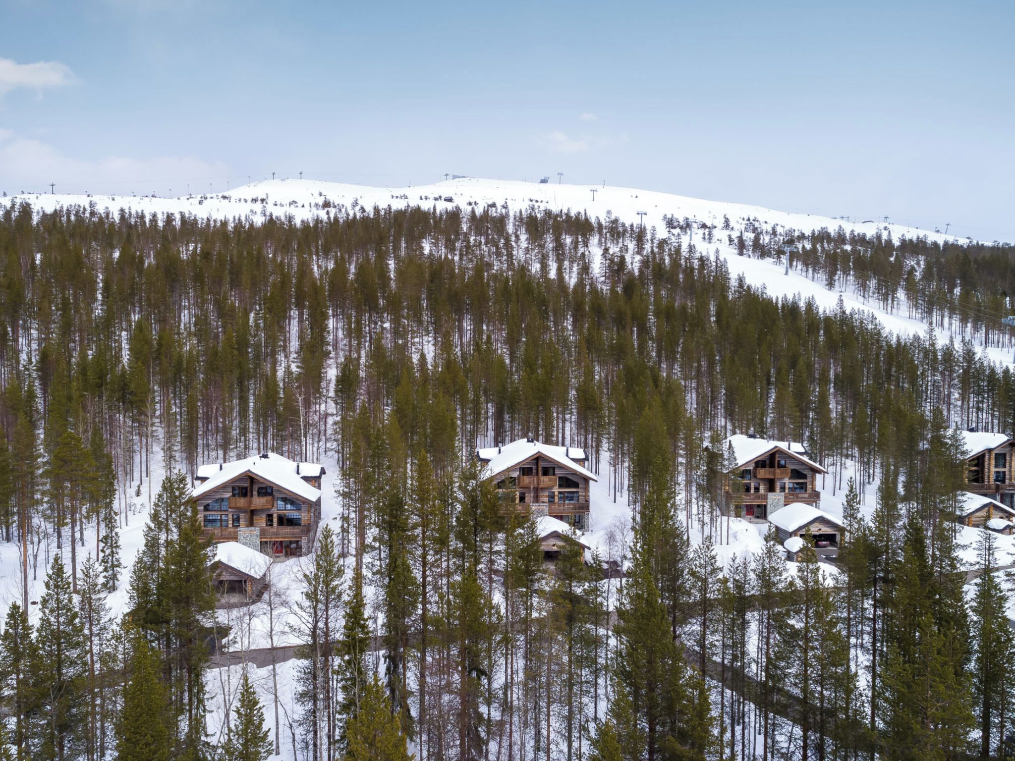 Un conjunto de cabañas de madera con techos nevados, rodeado por un bosque denso y una colina cubierta de nieve.
