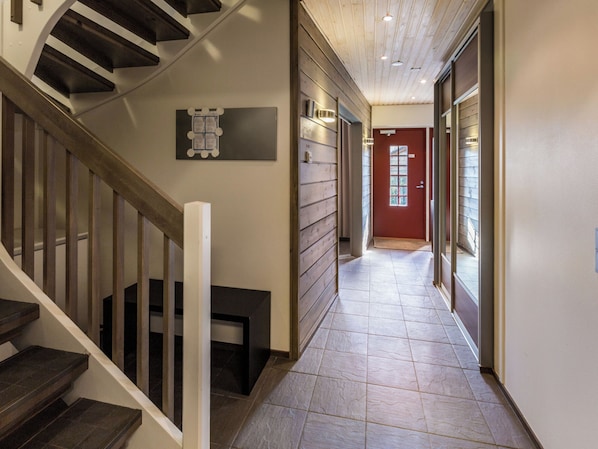 Un pasillo con una escalera de madera, una puerta roja y un piso de baldosas.