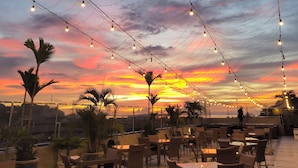 An outdoor seating area with tables and chairs, string lights overhead, and a sunset view. - Rylich Panorama Hotel (Sorong)