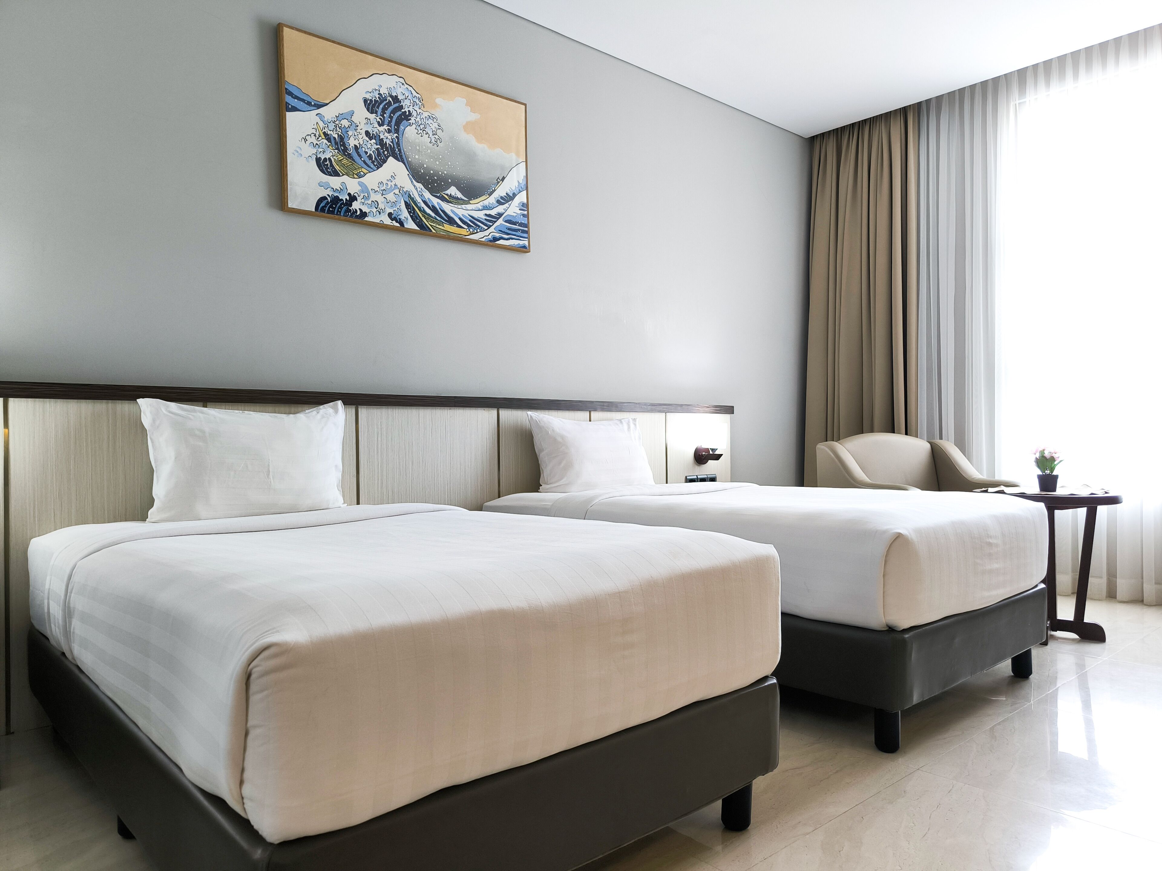 A hotel room with two beds, a painting on the wall, a chair, and a window with curtains.