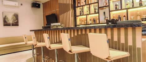 A bar area with a wooden panel wall, bar stools, and a shelf with various bottles.