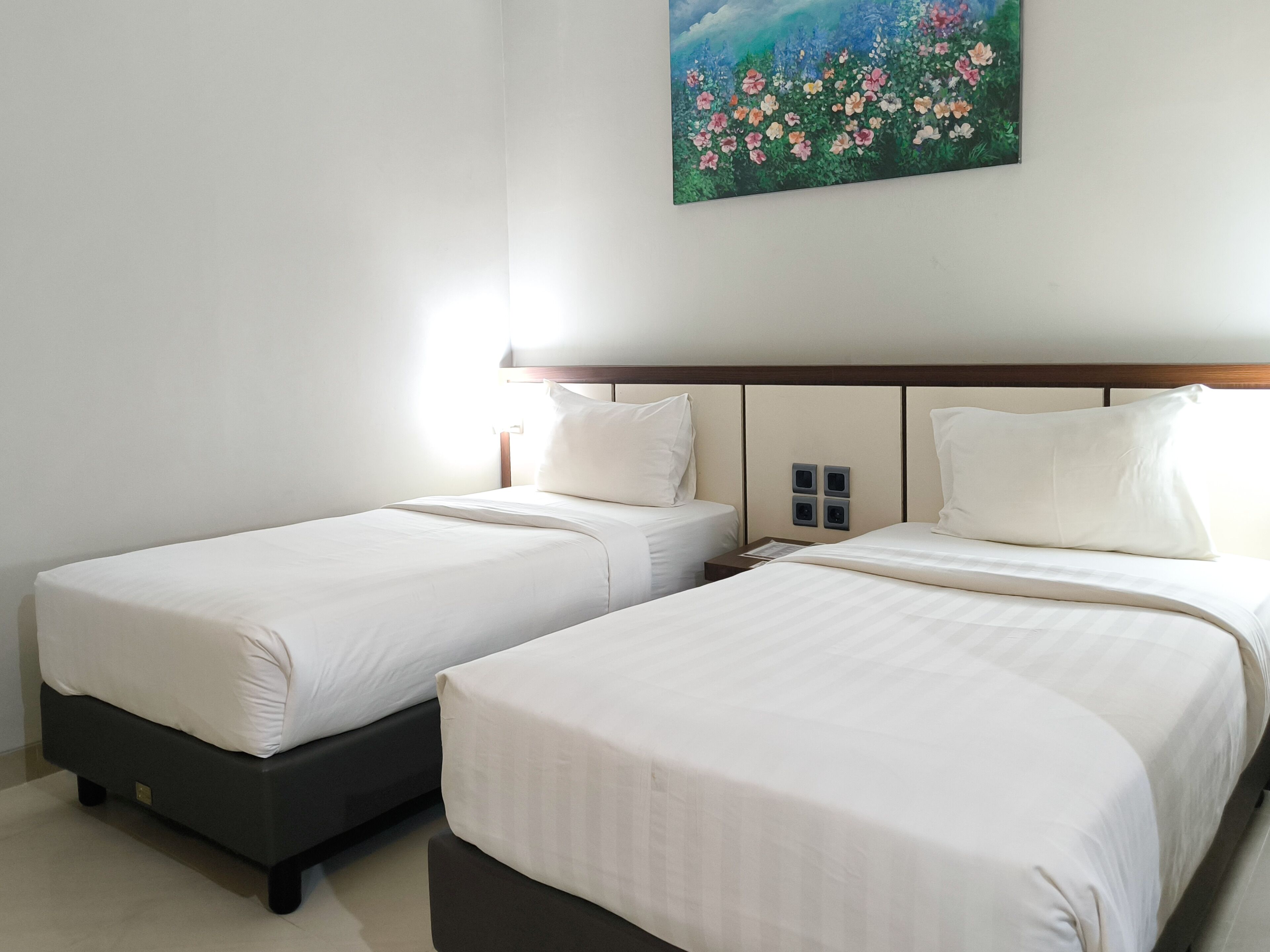 Habitación de hotel con dos camas individuales, cabecera de madera, un cuadro de flores en la pared y una mesita de noche con lámpara.